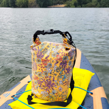 A multicolor floral dry bag with black straps sits on a bright yellow floatation pad on a paddleboard, with water in the background and black patterned sandals in the foreground.